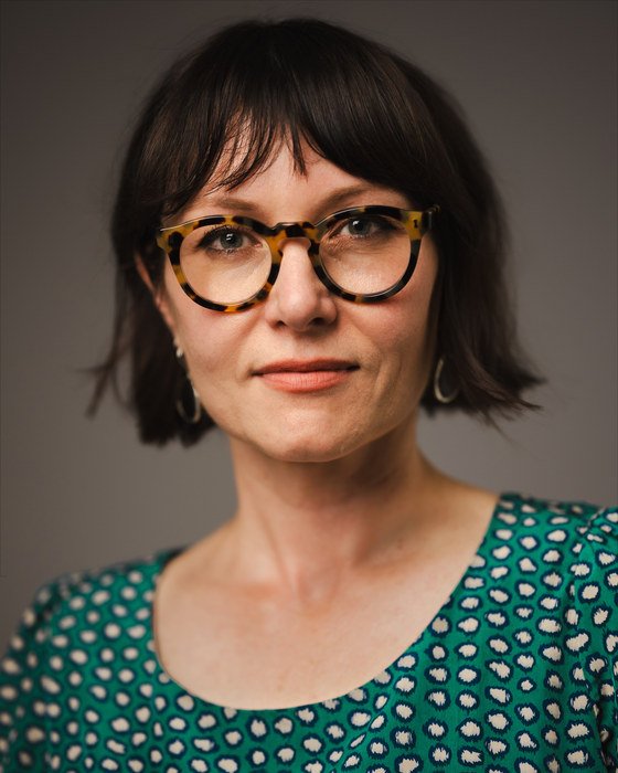 Leah Kardos in a green patterned dress against a grey-mauve studio backdrop, 2026